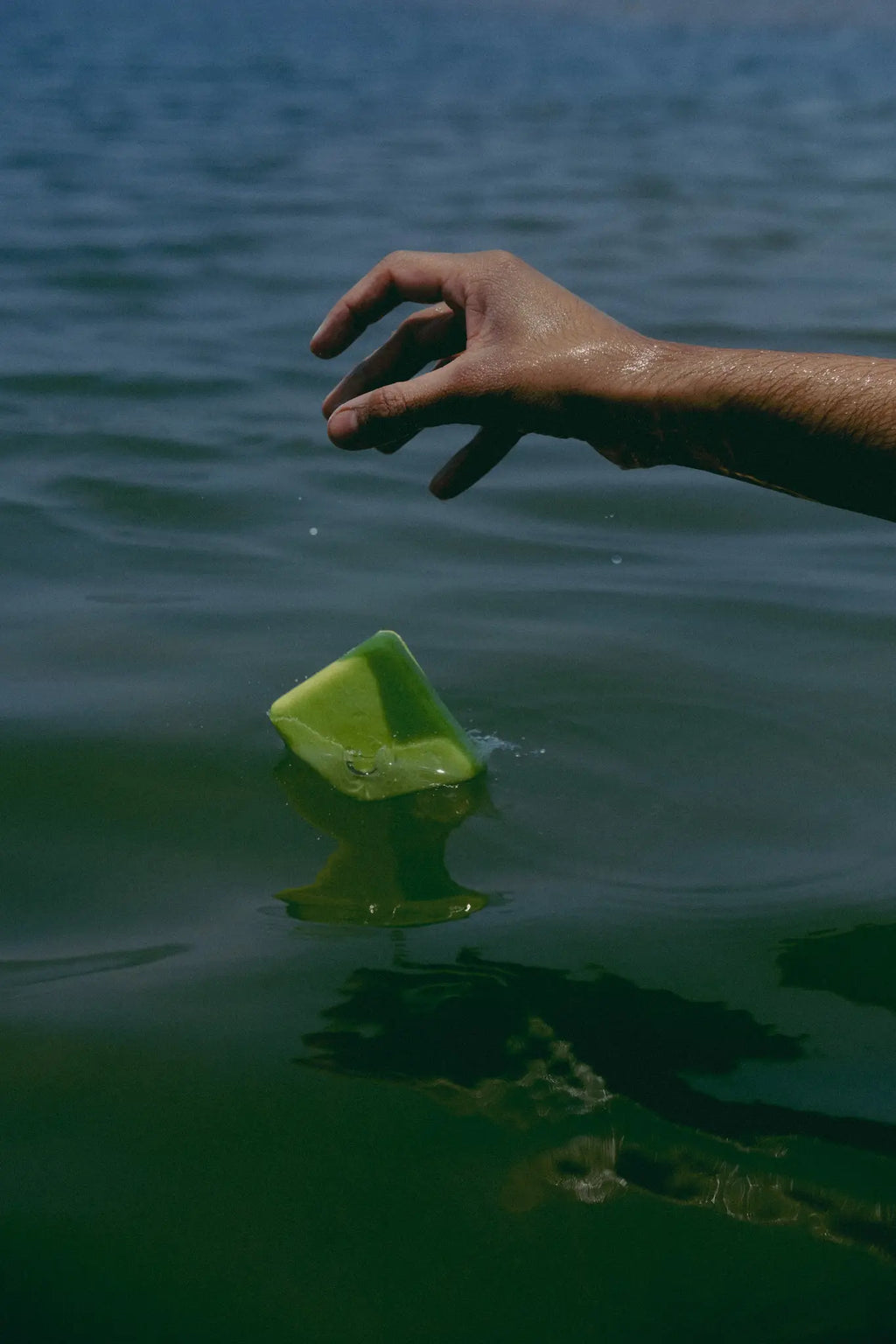 Hand reaching towards a green natural cold process soap floating on water